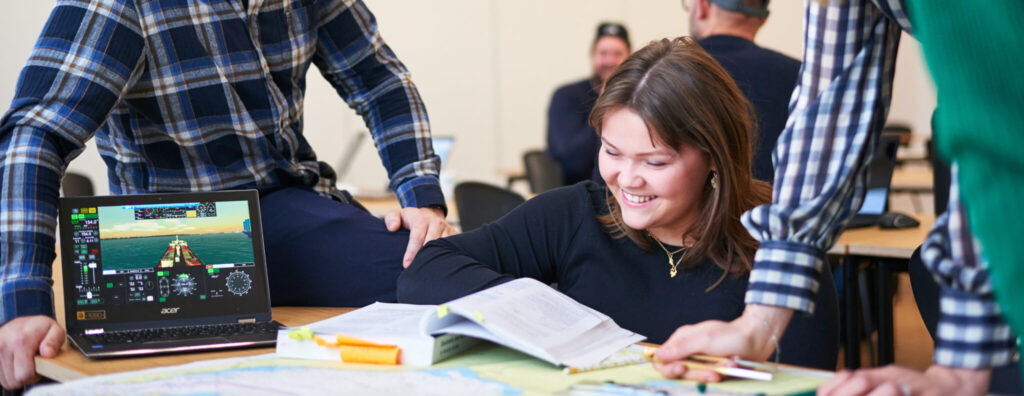 Skibsførerstuderende, der smiler og griner, mens de sidder lænet ind over søkort, studiebøger og skærm.
